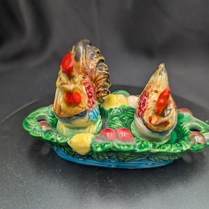 Puede incluir: Un juego de salero y pimentero de cerámica con figuras de gallo. Los gallos tienen crestas rojas y plumaje colorido. Se asientan sobre un plato verde en forma de hoja con detalles pintados, incluyendo acentos amarillos y rojos.