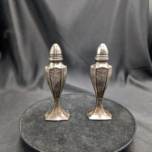 Può includere: Due saliere e pepaere antiche in argento. Ogni shaker ha un design a scudo decorativo e una parte superiore conica con fori. Gli shaker sono esposti su una superficie scura.