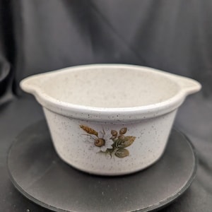 May include: A speckled, off-white ceramic baking dish with a floral design featuring a daisy and wheat stalks. The dish has two small handles. It is set on a dark surface. A charming addition to any kitchen.