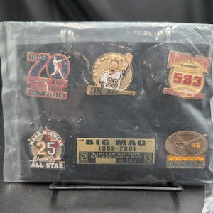 Puede incluir: Un set de colección de edición limitada con cinco pines de béisbol de Mark McGwire sobre un fondo negro dentro de una funda de plástico transparente. Los pines presentan varios diseños con el texto 1998 Home Run Season, 1986-2001, 583 Home Runs y Big Mac.