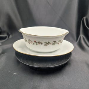 Może przedstawiać: Biała porcelanowa łódka do sosu z pasującym spodkiem. Łódka i spodek mają złote obramowanie i dekoracyjny wzór zielonych liści i czerwonych jagód. Łódka do sosu stoi na czarnym talerzu.