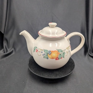 Könnte beinhalten: Weiße Keramik-Teekanne mit Deckel, Henkel und Ausguss. Die Teekanne ist mit einem Obst- und Beerenmuster in Orange, Grün und Blau verziert. Eine rote Zierleiste akzentuiert den Deckel. Die Teekanne steht auf einer schwarzen Oberfläche.