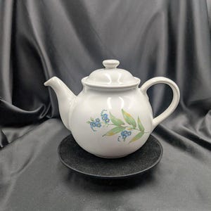 Könnte beinhalten: Weiße Keramik-Teekanne mit Deckel, Ausguss und Henkel. Die Teekanne hat ein Blumendesign mit blauen Blumen und grünen Blättern. Sie steht auf einem kleinen, runden, schwarzen Teller vor dunklem Hintergrund.