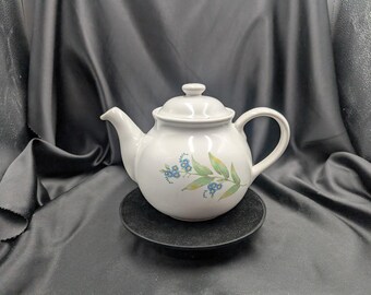 Op de afbeelding: Witte keramische theepot met deksel, tuit en handvat. De theepot heeft een bloemenontwerp met blauwe bloemen en groene bladeren. Hij staat op een klein, rond, zwart bordje tegen een donkere achtergrond.