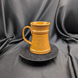 Puede incluir: Un jarro de cerveza de cerámica marrón con asa, sobre un plato negro. El jarro tiene las palabras "Alexander Keith's Fine Beers" y un emblema de ciervo. El jarro tiene un borde ancho y bandas decorativas.