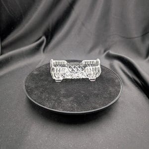 Può includere: Un piatto di vetro trasparente, rettangolare, con un motivo a diamante, appoggiato su una base circolare nera. Il piatto ha due scomparti ed è rialzato da piccoli piedini. Lo sfondo è un tessuto scuro drappeggiato.
