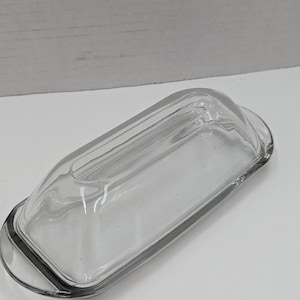 Op de afbeelding: Een heldere, rechthoekige glazen botervloot met een deksel. De schaal heeft afgeronde randen en een handvat aan elk uiteinde. Het deksel heeft een licht gebogen bovenkant met verticale ribbels. De schaal is leeg en staat op een wit oppervlak.