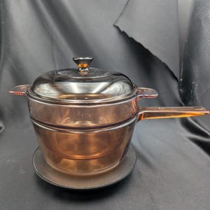 Pode incluir: Uma panela de vidro translúcido na cor âmbar com tampa a condizer. A panela apresenta um cabo longo e plano de um lado e uma pega mais pequena no lado oposto. A tampa tem um botão central e um padrão circular texturado no topo.