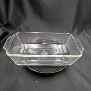Peut inclure: Un plat à four rectangulaire en verre transparent, avec des côtés légèrement évasés et de petites poignées. Le plat est vide et repose sur une surface sombre, mettant en valeur son design transparent. Idéal pour la cuisson et le service.