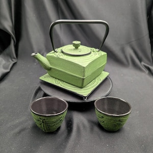 May include: A square, green cast iron teapot with a black handle and matching lid, resting on a square coaster. Two small, matching green cast iron tea cups with black interiors are positioned in front of the teapot on a circular black base.