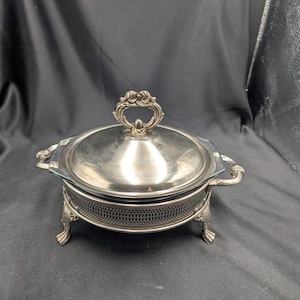 Op de afbeelding: Een zilverkleurige serveerschaal met een deksel en sierlijke handgrepen. De schaal heeft een glazen inzetstuk en staat op decoratieve voetjes. Het deksel heeft een gedetailleerde handgreep. De schaal is ontworpen voor het serveren van voedsel.