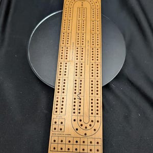 May include: A rectangular light brown wooden cribbage board featuring three parallel tracks of holes for scoring. The board includes printed markings such as "START," "ACME MADE IN CANADA," and "DESIGN REGISTERED - No. 14628" with a central letter S.