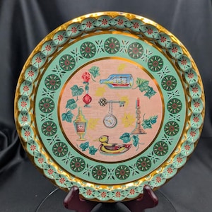May include: A circular vintage serving tray with a gold rim and a decorative edge. The tray has a light green and pink centre with illustrations of a bottle, key, clock, and duck. The rim has a repeating floral pattern.