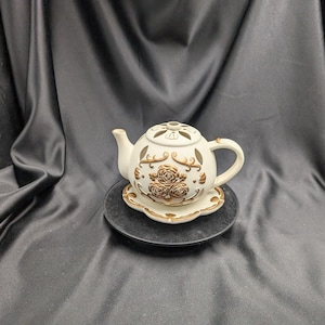 Puede incluir: Tetera de cerámica blanca con diseño floral dorado y base decorativa. La tetera tiene un diseño de corte y es perfecta para una fiesta de té o decoración del hogar.