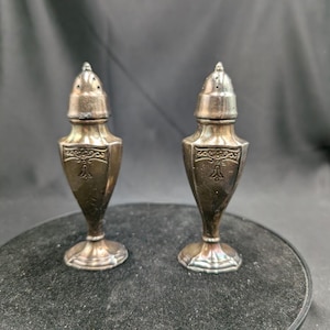 Può includere: Due saliere e pepiere antiche in argento. Ogni shaker ha un design a scudo decorativo e una parte superiore perforata. Gli shaker sono esposti su una superficie scura.