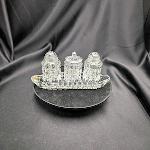 May include: A clear glass salt and pepper shaker set with a matching covered container, on a clear glass tray. The set is displayed on a black surface against a black background.