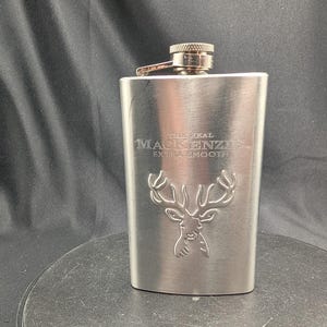 May include: A brushed stainless steel flask with a screw-top cap. The front features an embossed stag head design and the engraved text THE REAL MACKENZIE EXTRA SMOOTH. The metal container has a rectangular shape with rounded edges and a metallic finish.