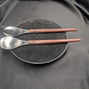 以下が含まれることがあります： ステンレス製のヘッドと木製のハンドルが付いたサラダサーバーのペア。片方のサーバーはスプーン型、もう片方は栓抜きのデザインです。ハンドルは濃い茶色で、黒い表面に置かれています。