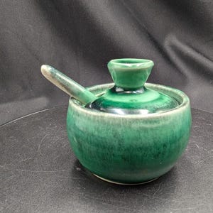 May include: A round ceramic sugar bowl with a glossy emerald green glaze featuring a lid with a central knob and a matching small spoon resting inside. The bowl has a smooth, curved body with darker green tones blending into lighter shades.
