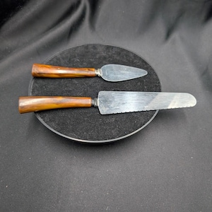 Può includere: Due utensili da portata con manici in legno e lame argentate poggiano su una superficie circolare nera. L'utensile più grande ha un bordo seghettato, mentre quello più piccolo è triangolare. I manici hanno una calda tonalità marrone.