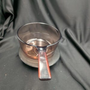 May include: A translucent, smoke-coloured glass saucepan with a single, integrated handle extending from the side. The circular vessel features a smooth, curved bowl and a flat-topped handle, showcasing a uniform dark tint throughout the glass material.