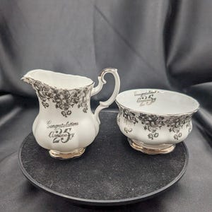 Może przedstawiać: Biały ceramiczny zestaw do śmietanki i cukiernicy. Zestaw posiada czarny kwiatowy wzór i złote wykończenia. Śmietanka i cukiernica mają napis "Congratulations 25 Anniversary" czarnym pismem.