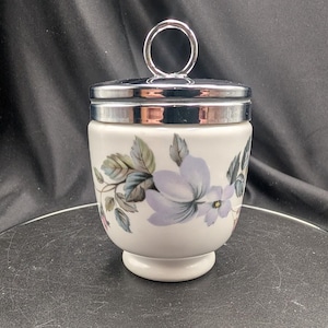 Könnte beinhalten: Ein weißes Keramikgefäß mit einem Blumenmuster aus hellvioletten Blüten und grünen Blättern. Das Gefäß hat einen silberfarbenen Metalldeckel mit einem runden Griff oben. Der Boden ist als Standfuß geformt und der Körper ist bauchig.