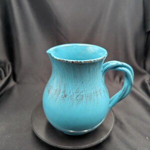 Peut inclure: Une cruche en céramique turquoise avec une anse torsadée et un petit bec verseur. La cruche a un corps arrondi et repose sur une assiette noire. L'émail a un aspect vieilli avec des accents plus foncés.