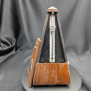 May include: A wooden metronome with a pyramid shape. The metronome has a dark wood finish with a lighter wood accent on the front. A silver pendulum and scale are visible inside. The metronome's door is open, revealing the interior.
