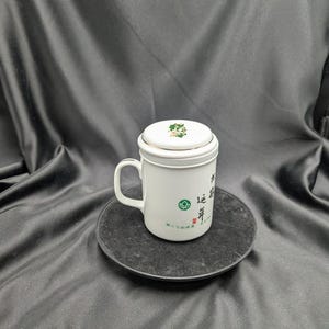 May include: White ceramic teacup with a lid and a green and black design. The cup has a handle and the lid has a small green leaf design. The cup is sitting on a black circular coaster.