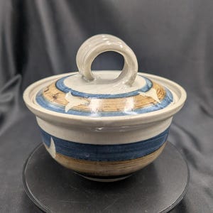 Puede incluir: Un cuenco de cerámica con tapa. El cuenco es blanco con rayas azules y beige y un asa curva. La tapa tiene un diseño azul y beige. El cuenco está sobre una superficie negra.