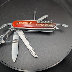 Puede incluir: Una navaja multiusos con mango rojo que presenta el texto Sheffield y Stainless Steel. La herramienta está totalmente desplegada, mostrando varios utensilios de metal plateado, incluyendo una hoja de sierra, una hoja de cuchillo y unas tijeras.