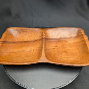 May include: A two-section wooden serving dish with a natural wood grain finish. The dish has a rectangular shape with gently curved edges. The interior of each section is slightly recessed. The dish is displayed on a black surface.