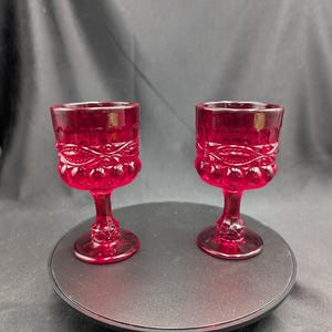 Op de afbeelding: Twee robijnrode kelkglazen met sierlijke details. De glazen hebben een voet en een decoratief patroon rond de kom. De kelkglazen zijn gemaakt van glas en zijn geschikt voor het serveren van dranken.