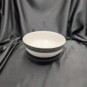 May include: A white ceramic bowl with a decorative band featuring a green, red, and gold diamond pattern. The bowl is round and sits on a dark surface, with a black background.