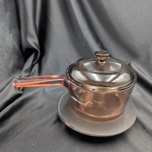 Peut inclure: Une casserole en verre ambré translucide avec un couvercle assorti et une longue poignée intégrée. Le couvercle présente un bouton circulaire et une texture légèrement nervurée autour du centre. Le corps en verre est lisse et s'affine vers la base.