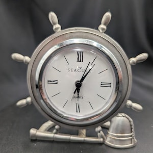 May include: A silver-colored nautical-themed clock shaped like a ship's wheel. The clock face is white with black Roman numerals and hands. The clock has the word "SEAGULL" printed on the face and is supported by an anchor and a ship's helm.