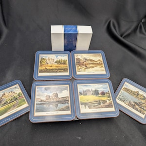 Könnte beinhalten: Set mit sechs quadratischen Untersetzern mit blauen Rändern, die jeweils eine andere Landschaftsszene eines Schlosses zeigen. Die Untersetzer sind um eine weiße Schachtel mit blauem Text angeordnet. Die Untersetzer bestehen aus einem haltbaren Material.