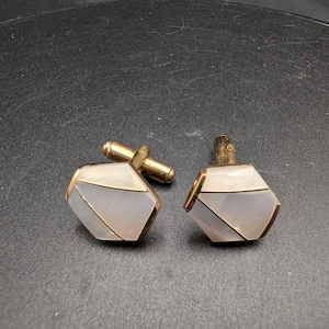 Puede incluir: Un par de gemelos con un diseño geométrico pentagonal, con incrustaciones de nácar blanco iridiscente engastadas en un marco de metal dorado. Cada gemelo cuenta con un mecanismo de cierre de barra basculante en la parte posterior.