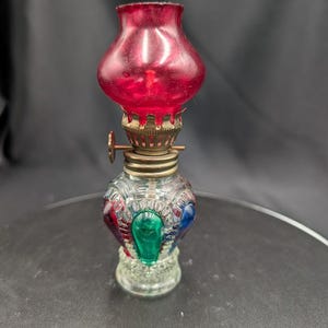 Op de afbeelding: Een kleine, decoratieve olielamp met een rode glazen kap en een heldere glazen voet. De voet is versierd met rode, groene en blauwe accenten. De lamp heeft een messingkleurig metalen mechanisme en een kleine knop voor de lont.