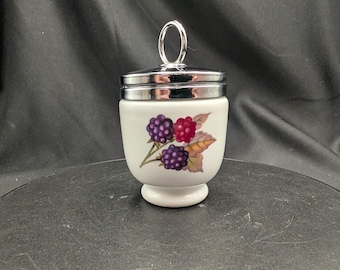 Pode incluir: Um cozedor de ovos de porcelana branca com tampa de metal prateado com uma pega em forma de anel. O corpo do recipiente é decorado com uma ilustração colorida de amoras roxas e vermelhas acompanhadas por folhas verdes e castanhas.