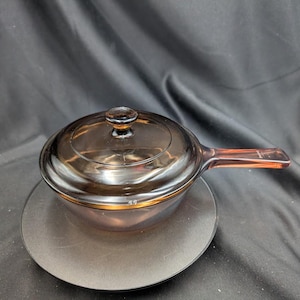 May include: A translucent amber-coloured glass saucepan with a matching circular lid. The cookware features a single integrated handle extending from the side and a small knob on the centre of the lid, crafted from heat-resistant glass material.