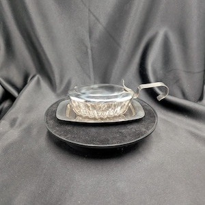 Peut inclure: Un petit plat de service argenté avec un couvercle et une poignée, posé sur un plateau rectangulaire. Le plat présente un motif texturé sur les côtés. Le plateau repose sur une base noire et circulaire sur fond sombre.