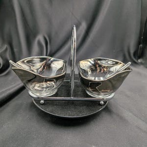 Może przedstawiać: Posrebrzany zestaw do przypraw z dwiema miseczkami i małymi łyżeczkami. Miseczki mają unikalny kształt i stoją na chromowanym stojaku z uchwytem. Zestaw przeznaczony jest do serwowania sosów, dipów lub innych małych porcji.