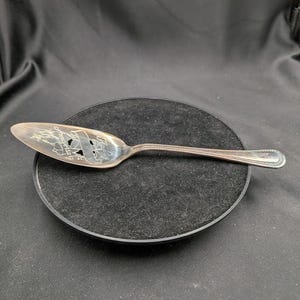 Puede incluir: Servidor de pastel plateado con un mango decorativo y un diseño calado detallado en la hoja. La hoja presenta el texto "Anniversary 1944" dentro de una pancarta y detalles florales. El servidor descansa sobre una superficie negra.