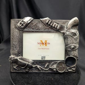 Peut inclure: Cadre photo en métal moulé argenté avec des ornements sur le thème de l'Ouest. Comprend un pistolet, une ceinture, des balles, un éperon et une montre. Le cadre peut contenir une photo de 14 cm x 23 cm et porte l'inscription "Marquis" et "Cast Metal Frame".