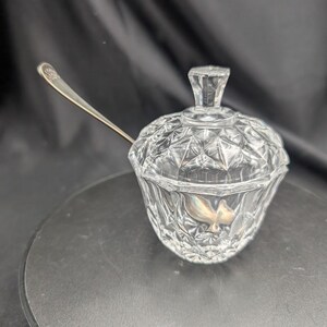 Peut inclure: Un sucrier en verre transparent orné d'un motif à facettes en diamant sur le corps et le couvercle, surmonté d'un bouton hexagonal. Une fine cuillère en métal argenté repose à l'intérieur, dépassant sur le côté du récipient.