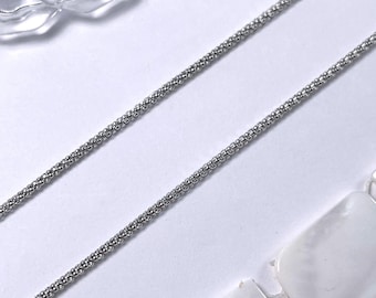 Könnte beinhalten: Eine silberne Kettenkette mit einem zarten, strukturierten Design. Die Kette liegt flach auf einer weißen Oberfläche.