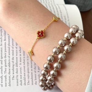 Puede incluir: Dos pulseras en una muñeca. Una pulsera es una cadena de oro con un dije de trébol rojo y pequeñas piedras transparentes. La otra pulsera es una doble hilera de perlas grandes de color marrón claro.