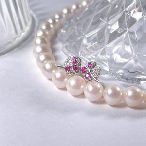 May include: A pair of silver earrings with pink gemstones and white diamonds. The earrings are on a white surface with a pearl necklace.